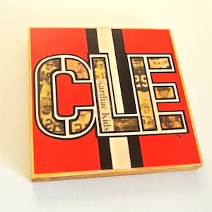 May include: A red, black, and white wooden sign with the word "CLE" in large, bold letters. The letters are made up of images of Cleveland Browns football players and newspaper clippings. The text "Cardiac Kids" is visible in the center of the sign.