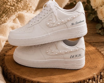 Könnte beinhalten: Ein Paar weiße Sneaker mit perlenbesetzten Swoosh-Logos an den Seiten. Der obere Schuh zeigt den Text Mr. Barahona, während der untere Schuh den Text Est 2026 in schwarzer Schreibschrift an den äußeren Fersenbereichen aufweist.
