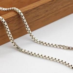 May include: A silver-toned box chain necklace featuring a series of interconnected square links that create a smooth, continuous, and flexible metallic strand. The chain has a polished finish that reflects light across its uniform, geometric structure.
