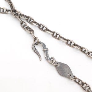 Op de afbeelding: Een zilverkleurige metalen ketting met ovale schakels en een verweerde afwerking. De ketting is voorzien van een decoratieve haaksluiting in de vorm van een vogel met gespreide vleugels en een klein, plat, ruitvormig metalen plaatje.