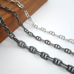 Puede incluir: Tres cadenas de metal con diseño de eslabones tipo ancla. Dos de las cadenas tienen un acabado oscuro y oxidado con textura visible, mientras que la tercera cadena es pulida y de color plateado brillante. Cada eslabón es de forma ovalada.