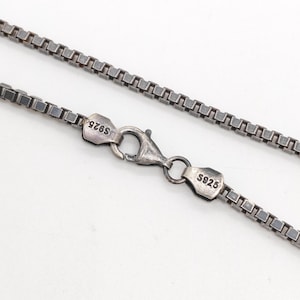 Può includere: Una catenina a maglia quadrata color argento con chiusura a moschettone. I componenti metallici alle estremità della catena riportano la scritta S925, che indica la composizione del materiale. La catena presenta maglie quadrate intrecciate.