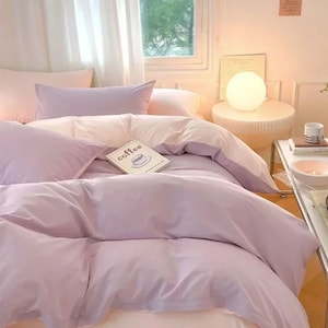 Può includere: Un letto con biancheria color viola chiaro, inclusi un copripiumino e cuscini. Un libro bianco con la parola coffee stampata sulla copertina poggia sul letto. Una lampada rotonda bianca si trova su un tavolino accanto al letto vicino a una finestra.