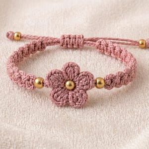 May include: A dusty pink macrame bracelet featuring a central crocheted flower with a gold-toned bead at its center. The band is crafted from knotted cord and includes two additional gold-toned beads flanking the flower, with an adjustable sliding knot closure.