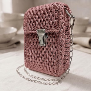 May include: A dusty rose crochet crossbody bag featuring a rectangular silhouette with a textured, woven pattern. The bag includes a silver-toned metal push-lock closure on the front flap and a detachable silver-toned metal chain shoulder strap.