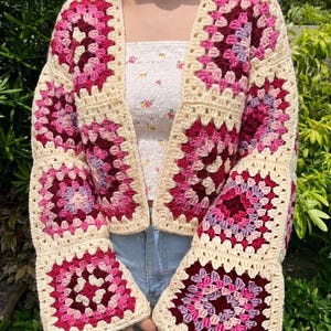 May include: A crochet cardigan constructed from square motifs featuring a cream-coloured border and a central pattern of concentric rings in shades of dark red, magenta, bright pink, and light purple, with long sleeves and an open front design.