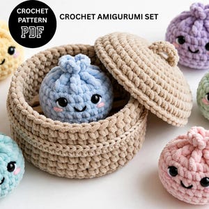 May include: A crochet amigurumi set featuring a beige yarn basket with a matching lid, containing a light blue plush dumpling character with black eyes and pink cheeks. Additional colorful plush dumplings in yellow, purple, green, and pink surround the basket.
