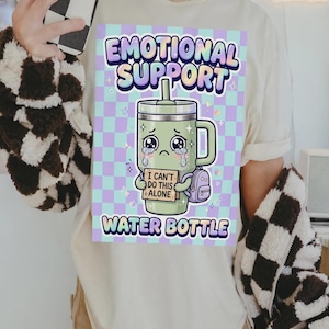 Puede incluir: Una camiseta de color crema con un gráfico de un vaso verde llorando con una pajita y una pequeña mochila morada. El vaso sostiene un cartel que dice "I CAN'T DO THIS ALONE". El texto "EMOTIONAL SUPPORT WATER BOTTLE" rodea la imagen.