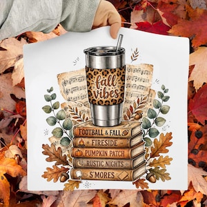 May include: A silver tumbler with a leopard print band featuring the text fall vibes, resting on a stack of five books labeled football & fall, fireside, pumpkin patch, rustic nights, and s'mores, surrounded by sheet music and autumn leaves.