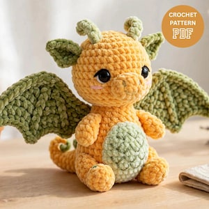 May include: A plush crochet dragon toy made of soft yellow yarn with sage green wings, horns, and a belly patch. The creature has large black safety eyes and pink stitched cheeks. A circular orange badge in the corner reads CROCHET PATTERN PDF.
