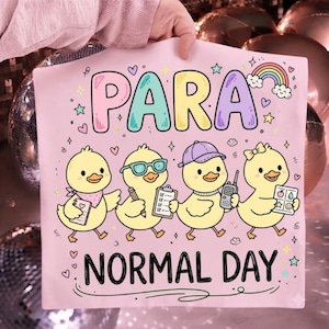 May include: A light pink t-shirt featuring the text PARA NORMAL DAY in colorful bubble letters. The design includes four yellow ducklings: one with a scarf and notebook, one with glasses and a clipboard, one with a cap and radio, and one with a bow and icon chart, all surrounded by small hearts, stars, and a rainbow.