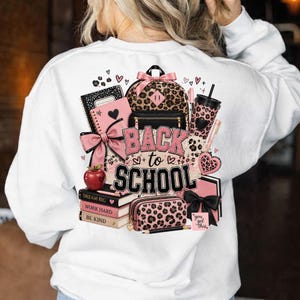 Pode incluir: Uma camisola branca com um gráfico nas costas com o texto BACK to SCHOOL. O design inclui uma mochila com padrão de leopardo, um caderno cor-de-rosa com um laço, uma maçã vermelha, livros empilhados com DREAM BIG, WORK HARD, BE KIND e um estojo.