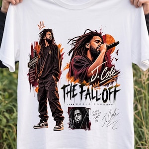 Puede incluir: Una camiseta blanca con un estampado gráfico del músico J. Cole. El diseño incluye varias imágenes del artista, el texto "J. Cole" y "The Fall Off World Tour", junto con salpicaduras de pintura abstractas en naranja y negro y coronas.