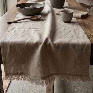 Può includere: Un lungo runner da tavola rettangolare in lino beige con bordi sfrangiati drappeggiato su un tavolo in legno. Il runner è arricchito da una ciotola in ceramica grigio scuro con pietre, un cucchiaio di legno, un vaso in pietra e una tazza.