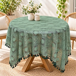 May include: A sage green round tablecloth featuring an intricate lace pattern with floral motifs and a grid design. The edges of the fabric are finished with a decorative fringe of small matching tassels that hang down around the perimeter.