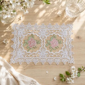 May include: A rectangular white lace doily featuring an intricate floral pattern with two central embroidered motifs of pink roses and green leaves. The lace border has a scalloped edge with repeating flower designs throughout the fabric.