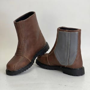 K&ouml;nnte beinhalten: Ein Paar wadenhohe Stiefel aus braunem Leder mit schwarzer Profilsohle. Ein Stiefel hat ein Seitenteil aus braunem Leder, w&auml;hrend der andere Stiefel ein kontrastierendes Seitenteil aus grauem Leder mit sichtbaren N&auml;hten aufweist.