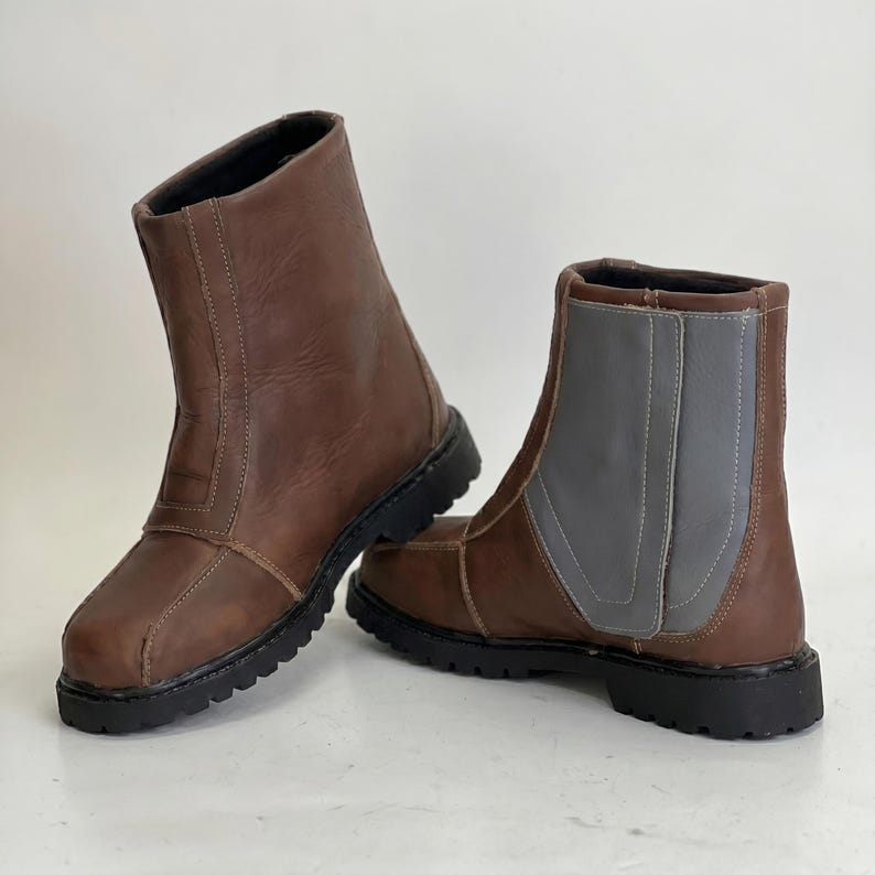 K&ouml;nnte beinhalten: Ein Paar wadenhohe Stiefel aus braunem Leder mit schwarzer Profilsohle. Ein Stiefel hat ein Seitenteil aus braunem Leder, w&auml;hrend der andere Stiefel ein kontrastierendes Seitenteil aus grauem Leder mit sichtbaren N&auml;hten aufweist.