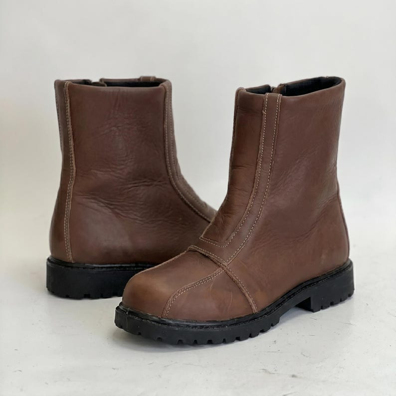 K&ouml;nnte beinhalten: Ein Paar wadenhohe braune Lederstiefel mit abgerundeter Zehenpartie, einer vertikalen Naht an der Vorderseite und einer dicken schwarzen Profilsohle. Die Stiefel haben eine glatte Oberfl&auml;che mit hellen Ziern&auml;hten und einem seitlichen Rei&szlig;verschluss.