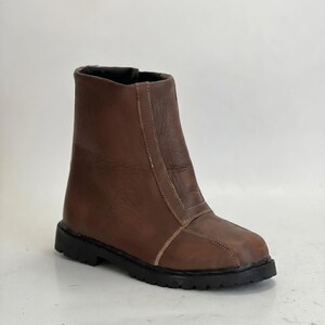 K&ouml;nnte beinhalten: Ein brauner Lederstiefel mit glatter Oberfl&auml;che, einer vertikalen Naht an der Vorderseite und einer dicken, schwarzen Gummisohle mit Profil. Der Stiefel hat eine abgerundete Zehenpartie, einen hohen Schaft und sichtbare N&auml;hte am Vorderteil.