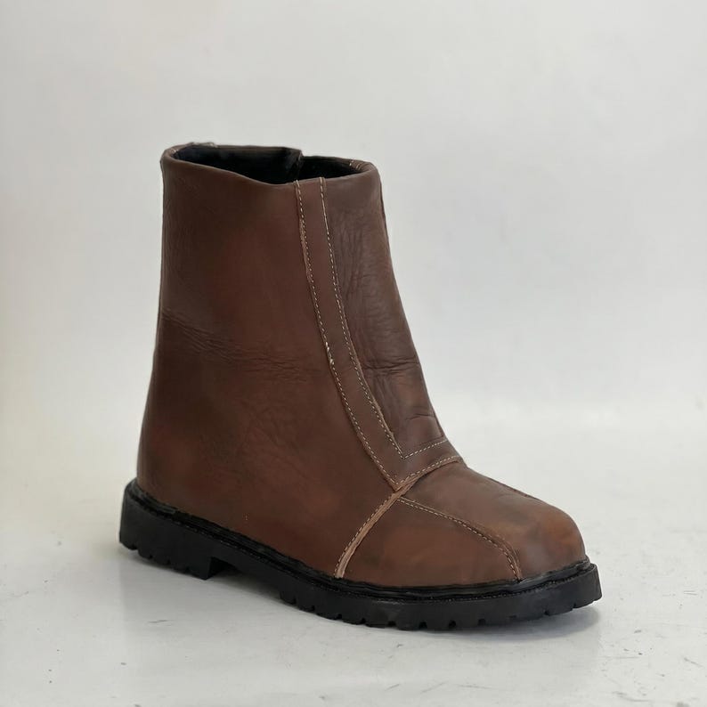 K&ouml;nnte beinhalten: Ein brauner Lederstiefel mit glatter Oberfl&auml;che, einer vertikalen Naht an der Vorderseite und einer dicken, schwarzen Gummisohle mit Profil. Der Stiefel hat eine abgerundete Zehenpartie, einen hohen Schaft und sichtbare N&auml;hte am Vorderteil.