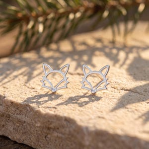 May include: A pair of silver-toned stud earrings shaped like the outline of a fox head. Each earring features a cut-out design with pointed ears and a defined snout, crafted from a polished metallic material that reflects light on its surface.