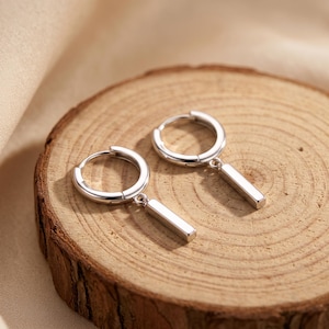 May include: A pair of silver-toned hoop earrings, each featuring a smooth, polished rectangular bar charm suspended from the bottom of the circular hoop. The earrings have a hinged closure mechanism and a reflective, metallic finish throughout.