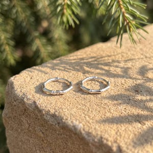 May include: A pair of silver hoop earrings featuring a segmented bamboo-inspired design. Each circular earring has a polished metallic finish with distinct nodes along the band, creating a textured appearance that mimics the look of natural bamboo stalks.