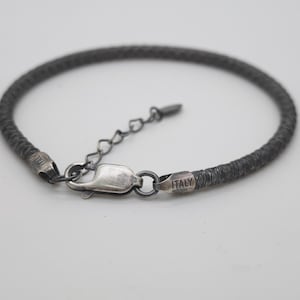 Puede incluir: Una pulsera de color gris oscuro con textura tejida, que cuenta con un cierre de mosquetón y una cadena de extensión ajustable. Los extremos metálicos llevan grabado el texto ITALY. El diseño consiste en un cordón fino y flexible con acabado.