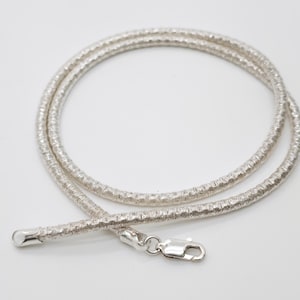 Puede incluir: Un collar de cadena de tono plateado con un diseño de eslabones redondeados y texturizados que recuerdan a una serpiente. El collar está enrollado en forma circular e incluye un cierre de mosquetón de metal pulido y un remate redondeado.