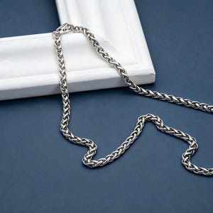 Op de afbeelding: Een zilverkleurige tarweketting met een doorlopend gevlochten schakelpatroon. De metalen ketting heeft een gepolijste, reflecterende afwerking en is voorzien van een kleine, veilige karabijnsluiting aan de bovenkant van de ketting.