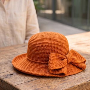 Peut inclure: Un chapeau au crochet orange brûlé doté d'une calotte arrondie et d'un large bord. Le chapeau est orné d'un grand nœud au crochet assorti fixé sur le côté du bord. La texture est uniforme, présentant un motif de mailles serrées et régulières.