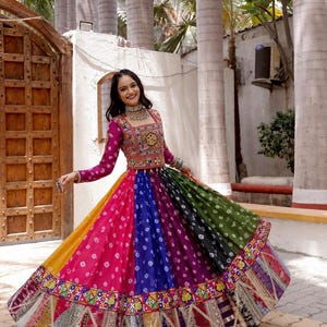 Puede incluir: Un lehenga choli multicolor que presenta una blusa de cuello cuadrado con bordados intrincados y espejos. La falda larga y acampanada muestra paneles verticales de tela amarilla, rosa, azul, morada y verde con un patrón de teñido anudado.