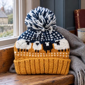 May include: A knit beanie featuring a mustard yellow ribbed cuff and a matching mustard yellow base. The upper section displays a pattern of white and black sheep against a dark navy blue background, topped with a large white and navy blue pom-pom.