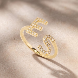 Puede incluir: Un anillo abierto de tono dorado que presenta las letras E y S, cada una incrustada con pequeñas piedras transparentes y brillantes. La banda es delgada y pulida, con las dos letras posicionadas en los extremos abiertos del anillo.