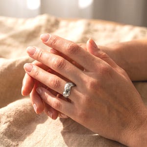 Puede incluir: Un anillo de plata con una superficie pulida, irregular y ondulada. La banda presenta un diseño grueso, orgánico y similar al metal fundido que envuelve el dedo, creando una apariencia metálica brillante y reflectante en toda la pieza.