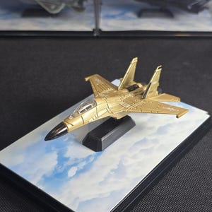 May include: A gold-colored miniature fighter jet model with a black nose cone and clear cockpit canopy. The aircraft is mounted on a black stand atop a rectangular base featuring a printed image of a blue sky with white clouds.