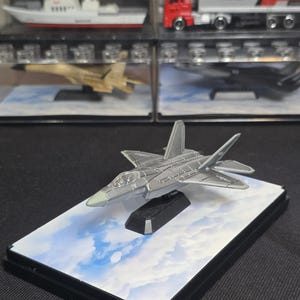May include: A die-cast metal model of a silver fighter jet mounted on a black display stand. The model is positioned on a rectangular base featuring a printed image of a blue sky with white clouds, showcasing detailed panel lines and a clear cockpit.