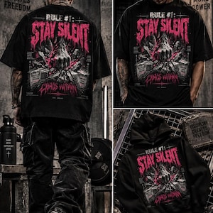 Könnte beinhalten: Ein schwarzes T-Shirt und ein schwarzer Kapuzenpullover mit einem Grafikdruck, der den Text "RULE #1: STAY SILENT" und "CHAOS WITHIN" in Pink zeigt. Das Design zeigt eine Faust, die eine Betonoberfläche durchbricht.