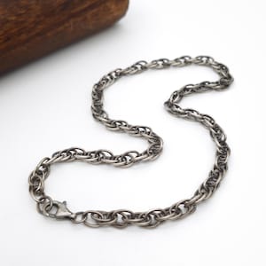 Può includere: Una collana a catena color argento scuro caratterizzata da una serie di maglie ovali intrecciate. Il metallo presenta una finitura leggermente ossidata o spazzolata, che gli conferisce un aspetto testurizzato. Un piccolo moschettone è fissato all'estremità.