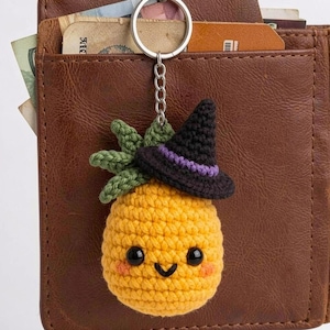 May include: A crochet pineapple keychain featuring a yellow textured body with black bead eyes, a small smile, and pink cheeks. The pineapple wears a black witch hat with a purple band and has green crocheted leaves attached to a silver metal ring.
