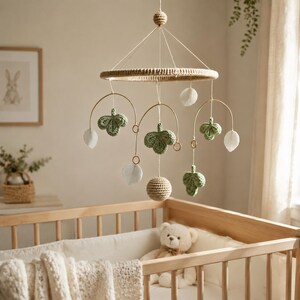 May include: A hanging nursery mobile featuring a circular frame wrapped in beige yarn. Suspended from the frame are three gold-toned metal arches, each adorned with crocheted green clover-like shapes, white felt leaves, and a central crocheted sphere.