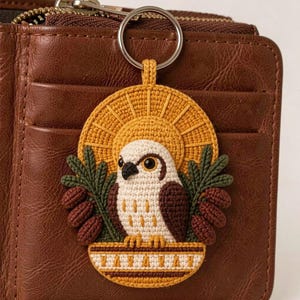 May include: A crocheted keyring featuring a white and brown bird perched in a decorative base, framed by a yellow sunburst and green pine branches with brown cones. The base includes a repeating geometric pattern in white, brown, and yellow.