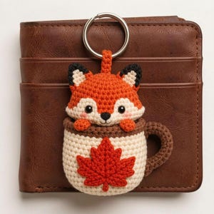 May include: A crochet keychain featuring a small orange and white fox peeking out of a cream-colored mug. The mug is decorated with a red maple leaf and has a brown handle. The keychain is attached to a silver metal ring and rests on a brown wallet.