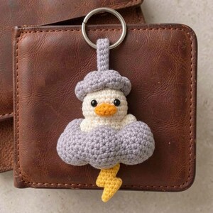 May include: A crochet keychain featuring a small white duck with an orange beak, nestled inside a light purple cloud. A yellow lightning bolt hangs from the bottom of the cloud, and a silver metal ring is attached to the top of the cloud.