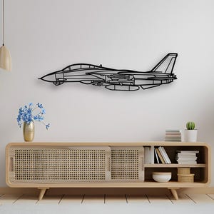 May include: A black metal wall art piece shaped like a fighter jet in profile. The design features the distinct silhouette of a military aircraft with a cockpit, wings, and tail fin, rendered in a clean, minimalist line-art style.