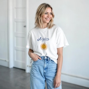 Puede incluir: Una camiseta blanca de manga corta con la palabra Argentina escrita en letra cursiva azul sobre un gráfico de sol amarillo con cara, combinada con unos vaqueros de mezclilla azul claro de talle alto y cierre de botones.