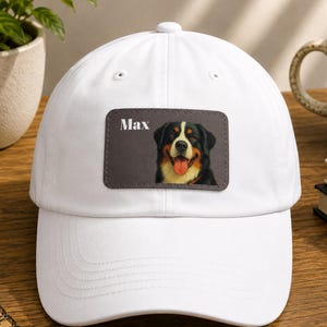 May include: A white baseball cap featuring a rectangular patch on the front. The patch displays a portrait of a Bernese Mountain Dog with its tongue out and the name Max printed in white text to the left of the dog's head on a dark background.