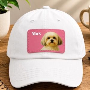 Può includere: Un cappellino da baseball bianco con una toppa rettangolare sul davanti. La toppa ha uno sfondo rosa con la grafica di un cane dal pelo chiaro e il testo bianco Max stampato nell'angolo in alto a sinistra della toppa.