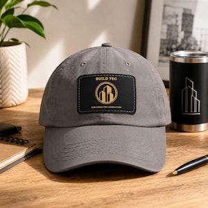 May include: A grey baseball cap featuring a rectangular black patch on the front. The patch displays a gold circular logo with building silhouettes and the text BUILD TEC and BUILDINGS FOR GENERATION in gold lettering with visible stitching.